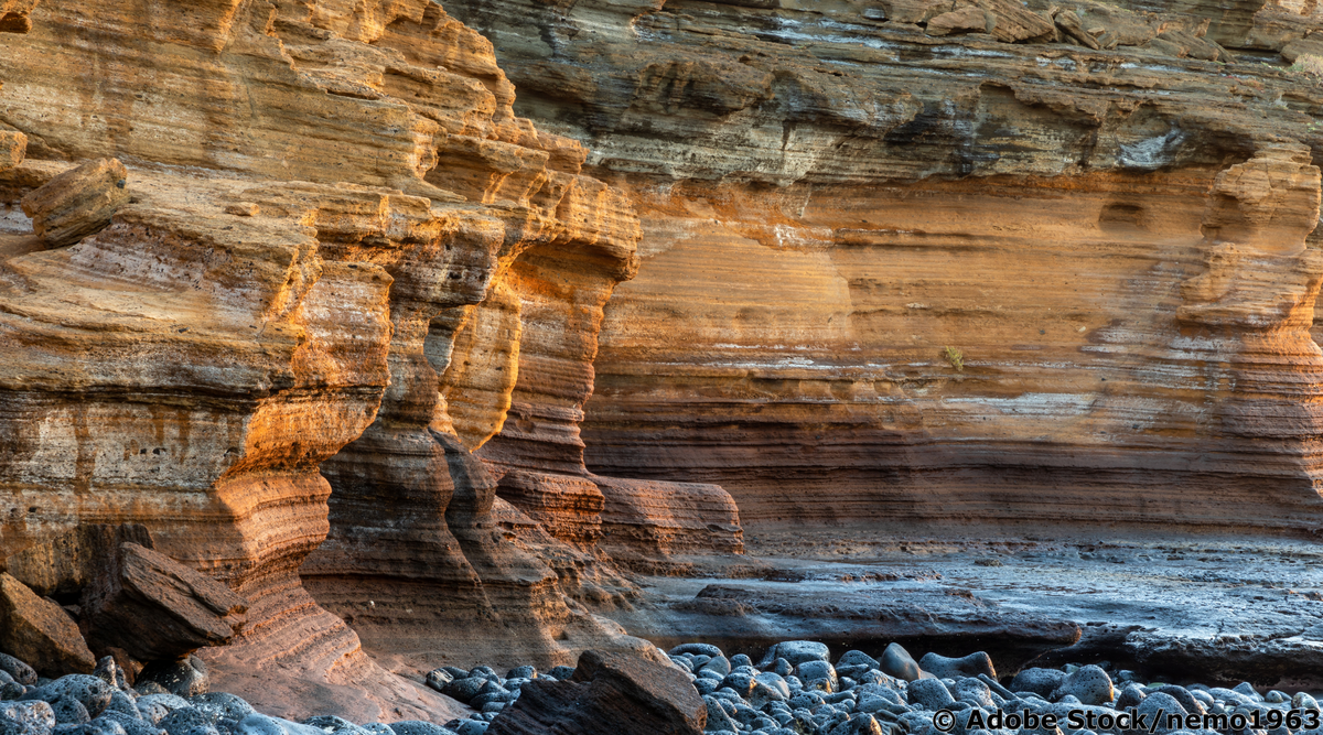 Geschichtete Sandsteinklippe am Meer mit horizontalen Sedimentschichten und Kieselstrand.