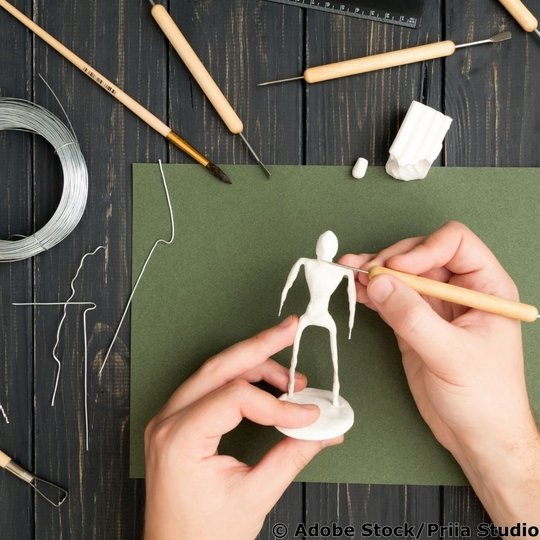 Hände modellieren eine schlanke Drahtfigur mit weißer Modelliermasse; daneben liegen Draht und Werkzeuge auf einem Holztisch.
