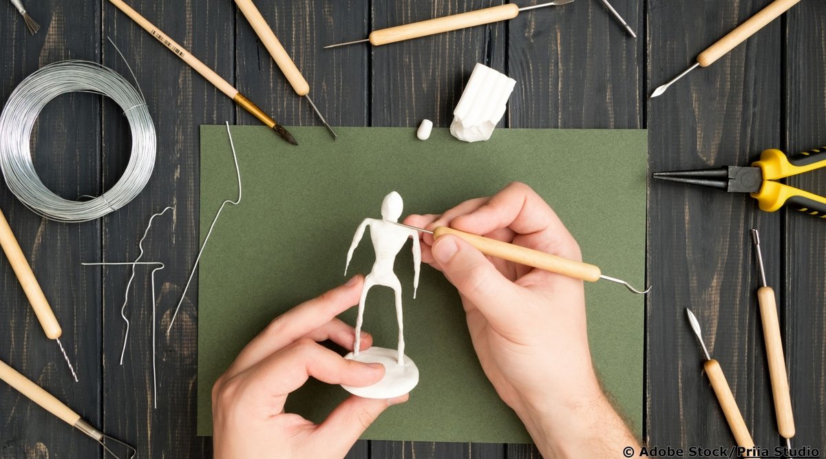 Hände modellieren eine schlanke Drahtfigur mit weißer Modelliermasse; daneben liegen Draht und Werkzeuge auf einem Holztisch.