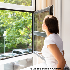 Junge Frau in weißem T-Shirt vor einem offenen Fenster, mit Blick auf grüne Bäume und einen Parkplatz