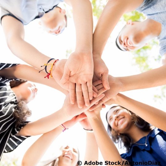 Das Bild zeigt eine Gruppe junger Menschen, die im Kreis stehen und ihre Hände in der Mitte übereinander legen, aufgenommen aus der Perspektive von unten nach oben.