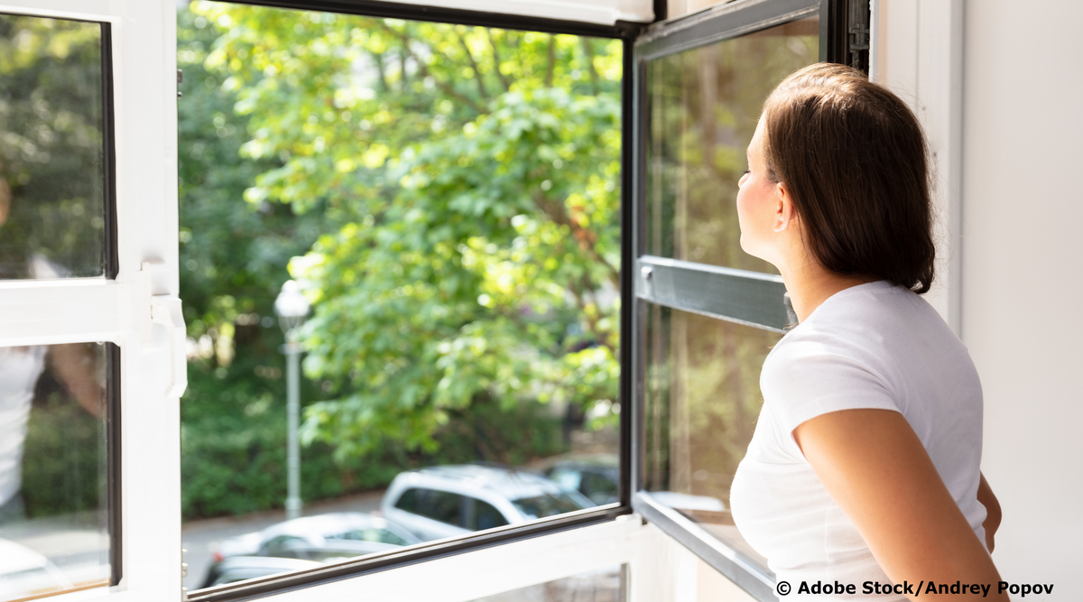Junge Frau in weißem T-Shirt vor einem offenen Fenster, mit Blick auf grüne Bäume und einen Parkplatz