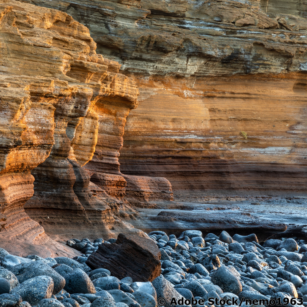 Geschichtete Sandsteinklippe am Meer mit horizontalen Sedimentschichten und Kieselstrand.